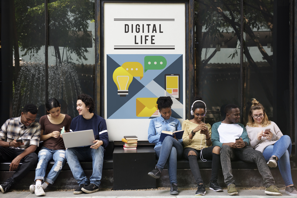 A licensed shutter stock image of a large sign with he title Digital Life and 7 teens sitting with different technology devices on benches in front of it.